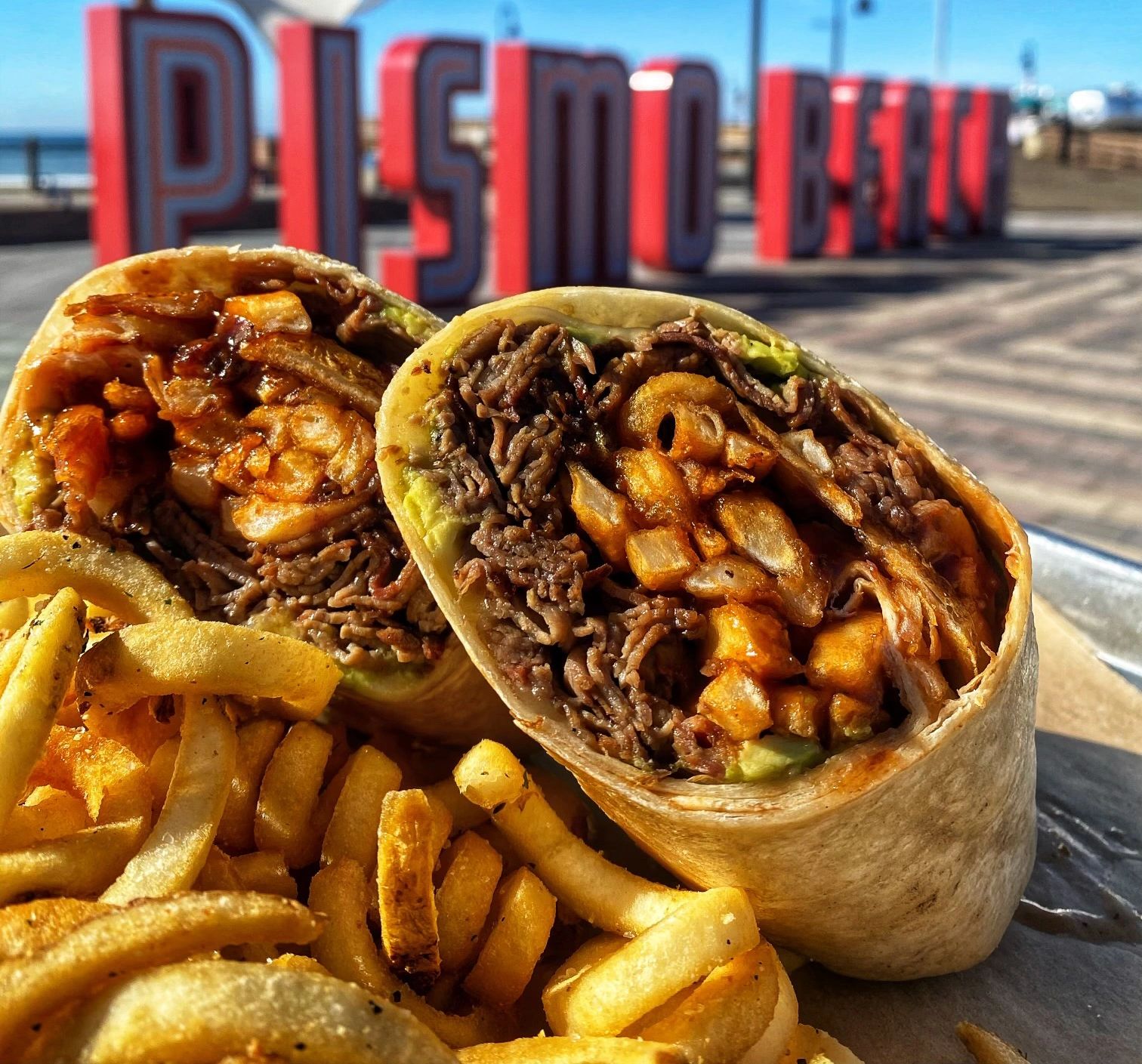 Pismo Wrap with tri tip, bbq, fries, cheese, and avocado in front of the Pismo Beach sign.