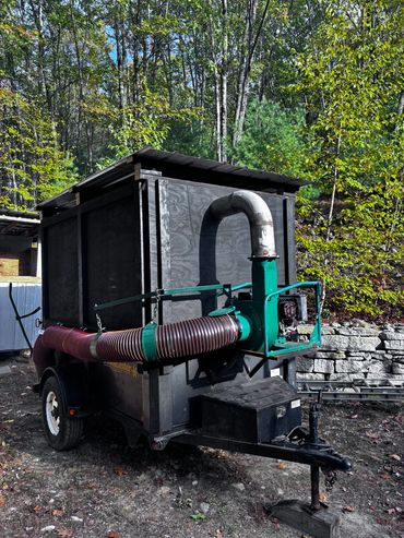 A leaf vacuum trailer with large hose and engine, parked near trees and a stone wall.