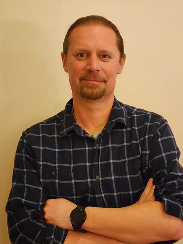 Man with a goatee wearing a plaid shirt and a smartwatch, smiling confidently.