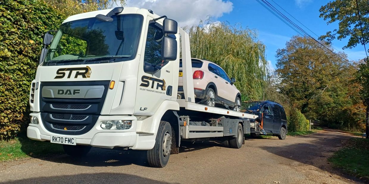 A white STR DAF recovery truck carrying a beige car on a sunny road for vehicle transport services and emergency vehicle recovery