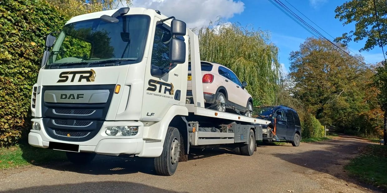 A white STR DAF recovery truck carrying a beige car on a sunny road for vehicle transport services and emergency vehicle recovery