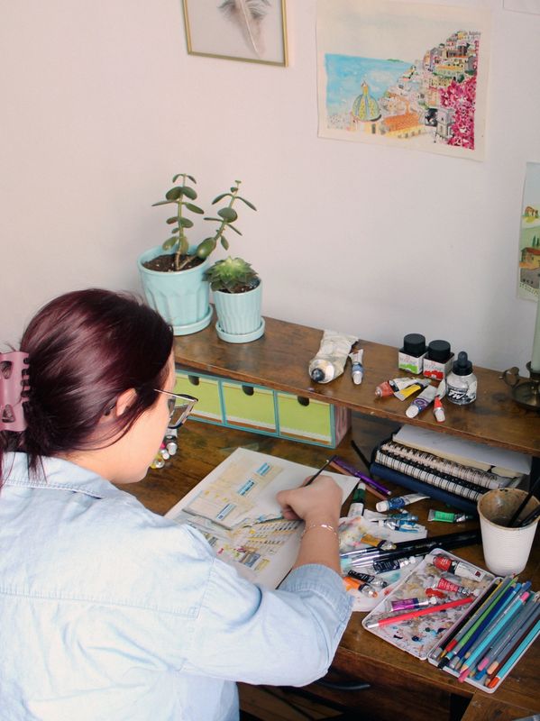 A person painting at a cluttered wooden desk with plants and art supplies.