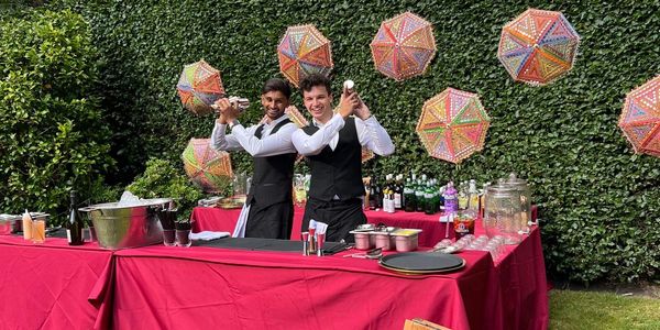 Two bartenders shaking cocktails at an outdoor bar with colorful decorations.