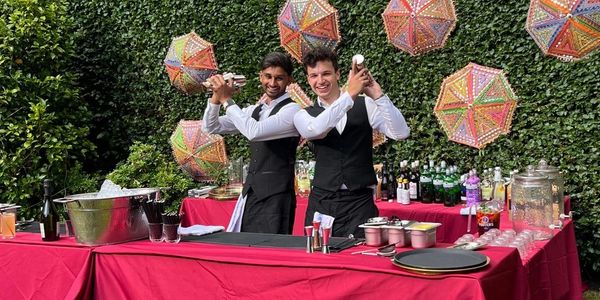 Two bartenders shaking cocktails at an outdoor bar with colorful decorations.