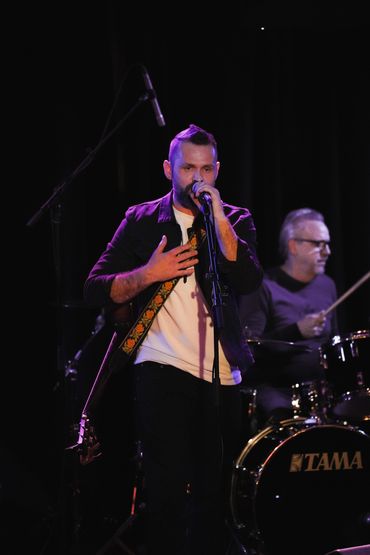Male singer passionately performing on stage with a drummer in the background.