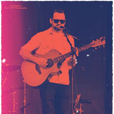 Man with sunglasses plays guitar and sings on stage with a microphone.