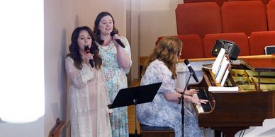 Three women performing music with piano and microphones in a church setting.