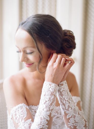 Bride with intricate lace sleeves adjusting her earring, smiling gently.