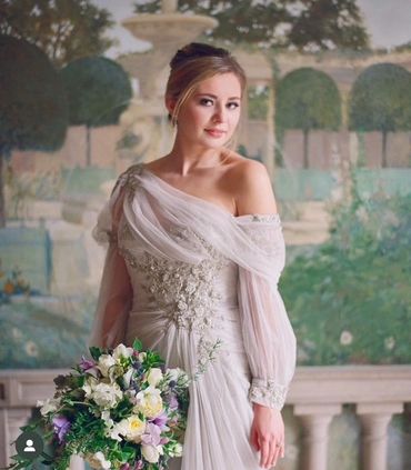 Bride in an elegant off-shoulder gown holding a bouquet against a garden mural.