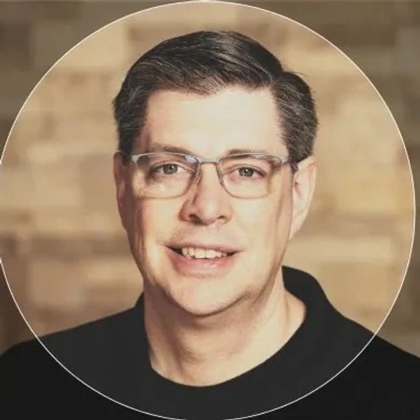 Smiling man with glasses in front of a blurred brick wall.