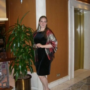 Woman in black dress posing indoors near a plant.