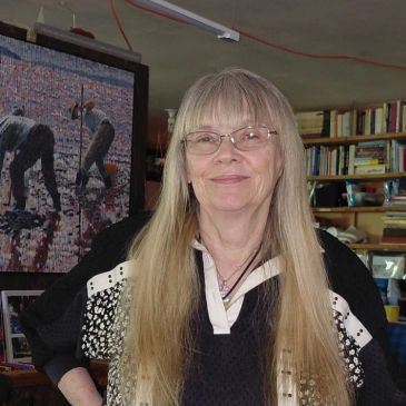Smiling woman with long blonde hair and glasses standing indoors.