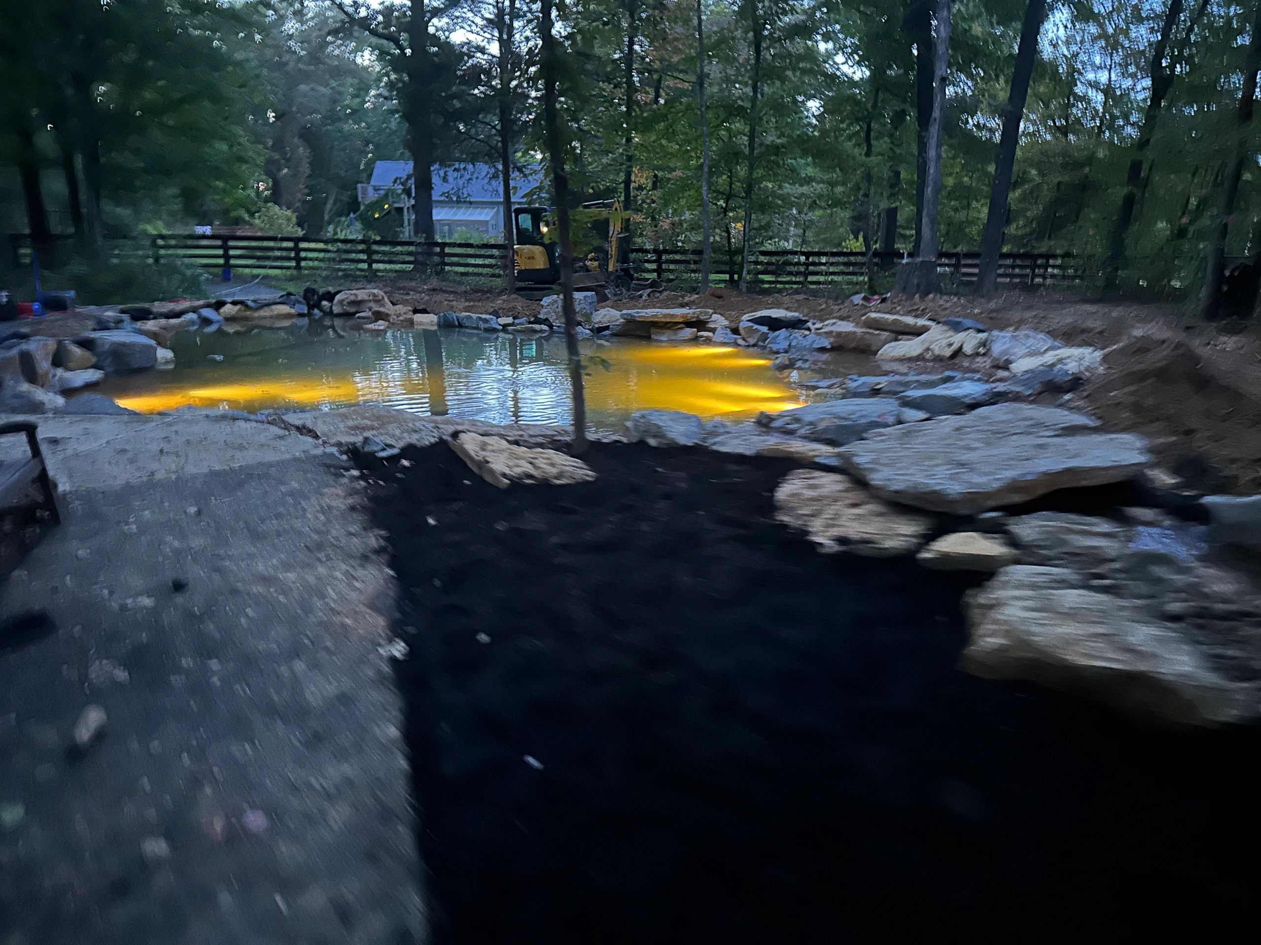 Backyard pond with yellow lights and excavator at dusk.