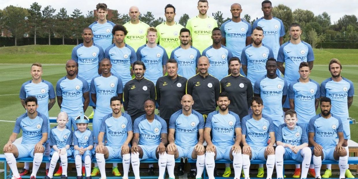 Manchester City football team posing in their sky blue kits on a training ground.