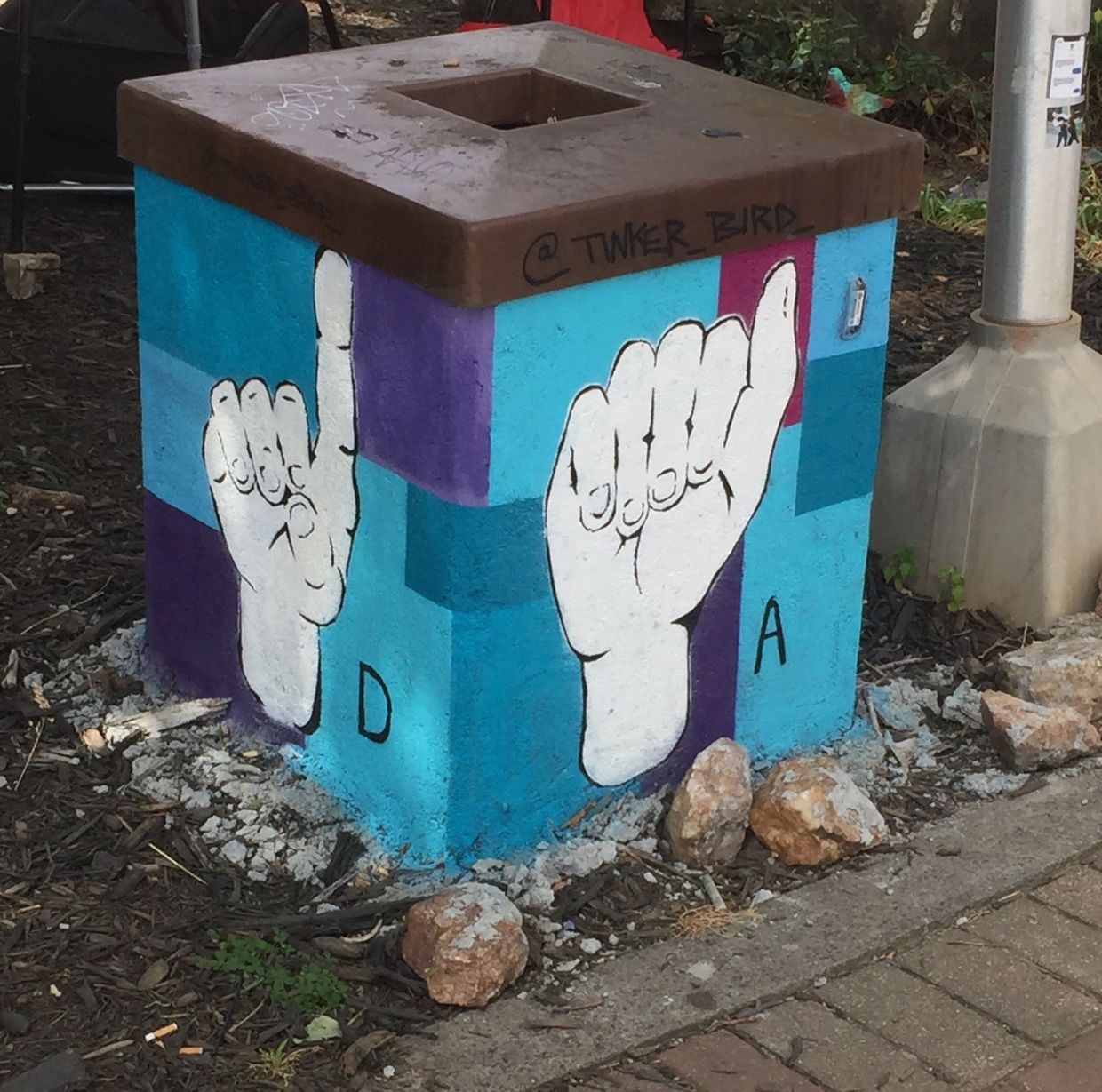 Large public trash can painted with a blue background and white hands signing the letters D and A