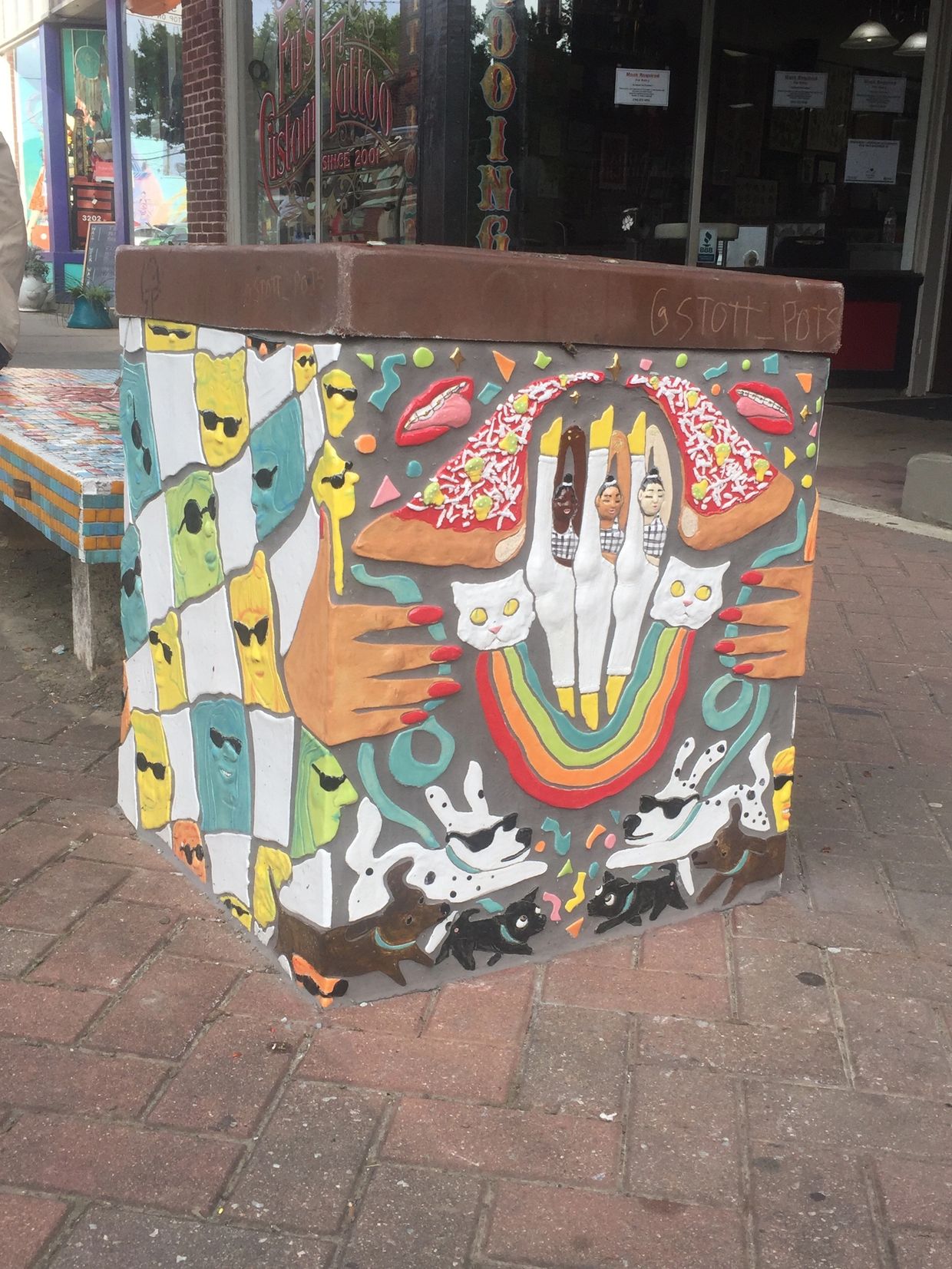 Public trash container covered in ceramic tile mosaic with dancing figures, dogs, cats, and pizza.