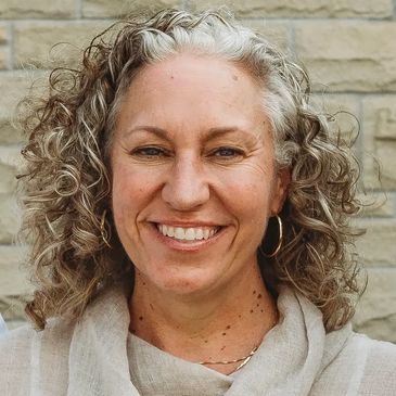 Smiling woman with curly gray hair in front of a stone wall.