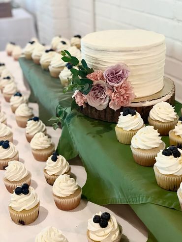 White wedding cake with pink and purple flowers and blueberry and vanilla cupcakes surrounding it