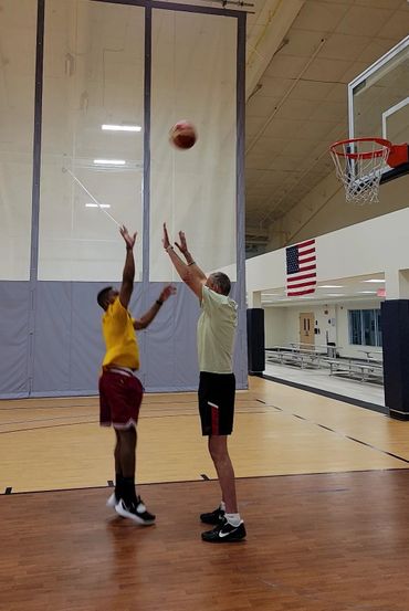 Two men playing basketball indoors, one shooting and the other defending.