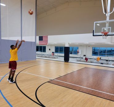 Man shooting a basketball indoors on a court with American flag in background.