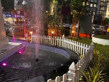 Nighttime fountain surrounded by a white picket fence and greenery in an urban setting.