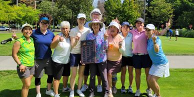 Group of women golfers posing with a plaque on a sunny golf course.