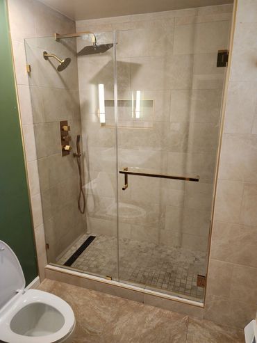 Modern beige tiled shower with glass door and gold fixtures beside a white toilet.
