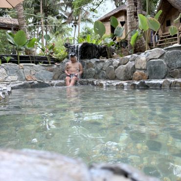 Person relaxing in a tropical pool with lush greenery and stone walls.