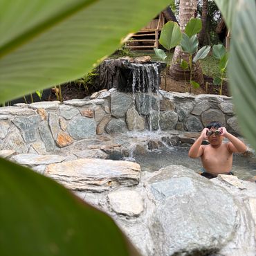 A child wearing goggles in a stone pool with a waterfall, surrounded by greenery.