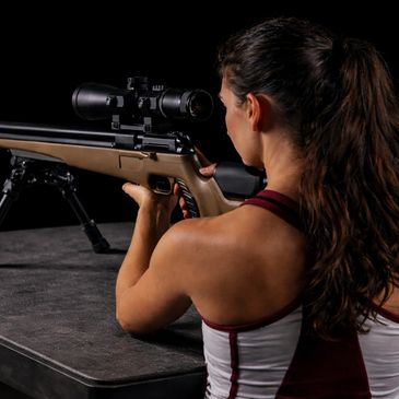 A woman aiming a rifle with a scope on a table, focused and ready.