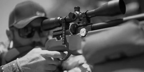 Man aiming a scoped rifle, wearing sunglasses and a cap.