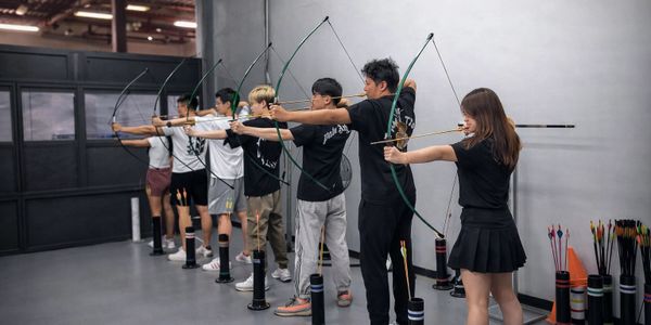 A group of people practicing archery indoors, aiming their bows in unison.