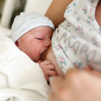 Newborn baby breastfeeding, wrapped in a white blanket and wearing a hat.