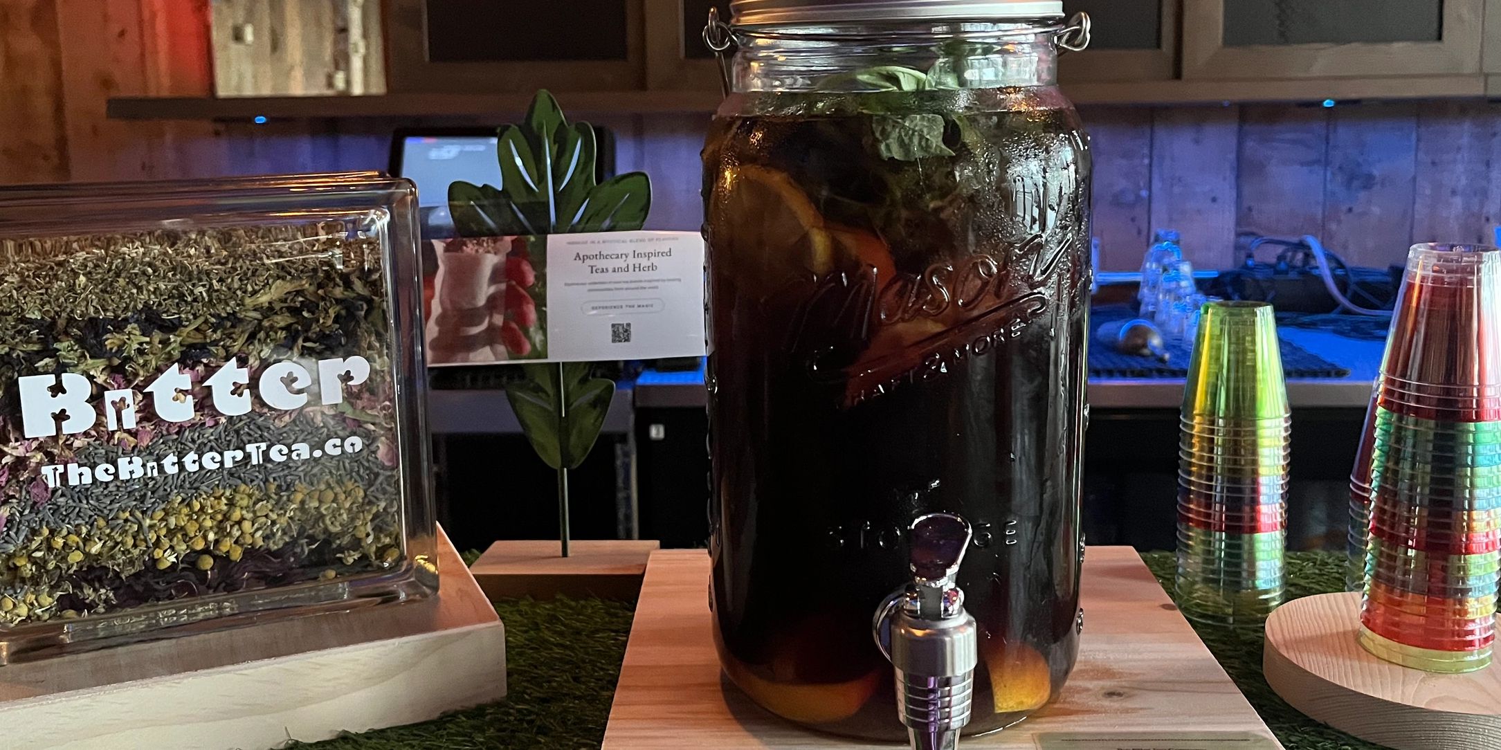 Large mason jar with a pouring spout on wooden platform
Next to a glass square sign with “Bitter”.