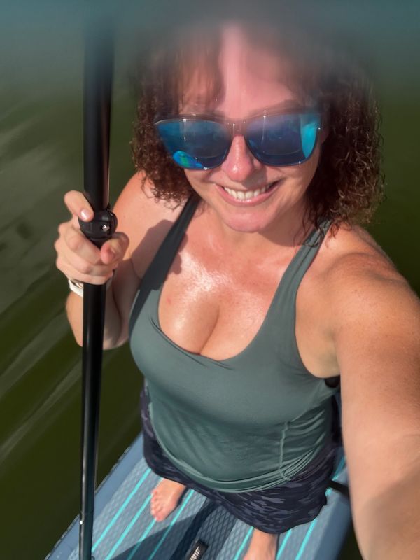 Woman paddleboarding on calm water, smiling and wearing sunglasses.