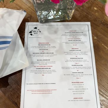 A restaurant menu on a wooden table with a flower vase and napkin nearby.
