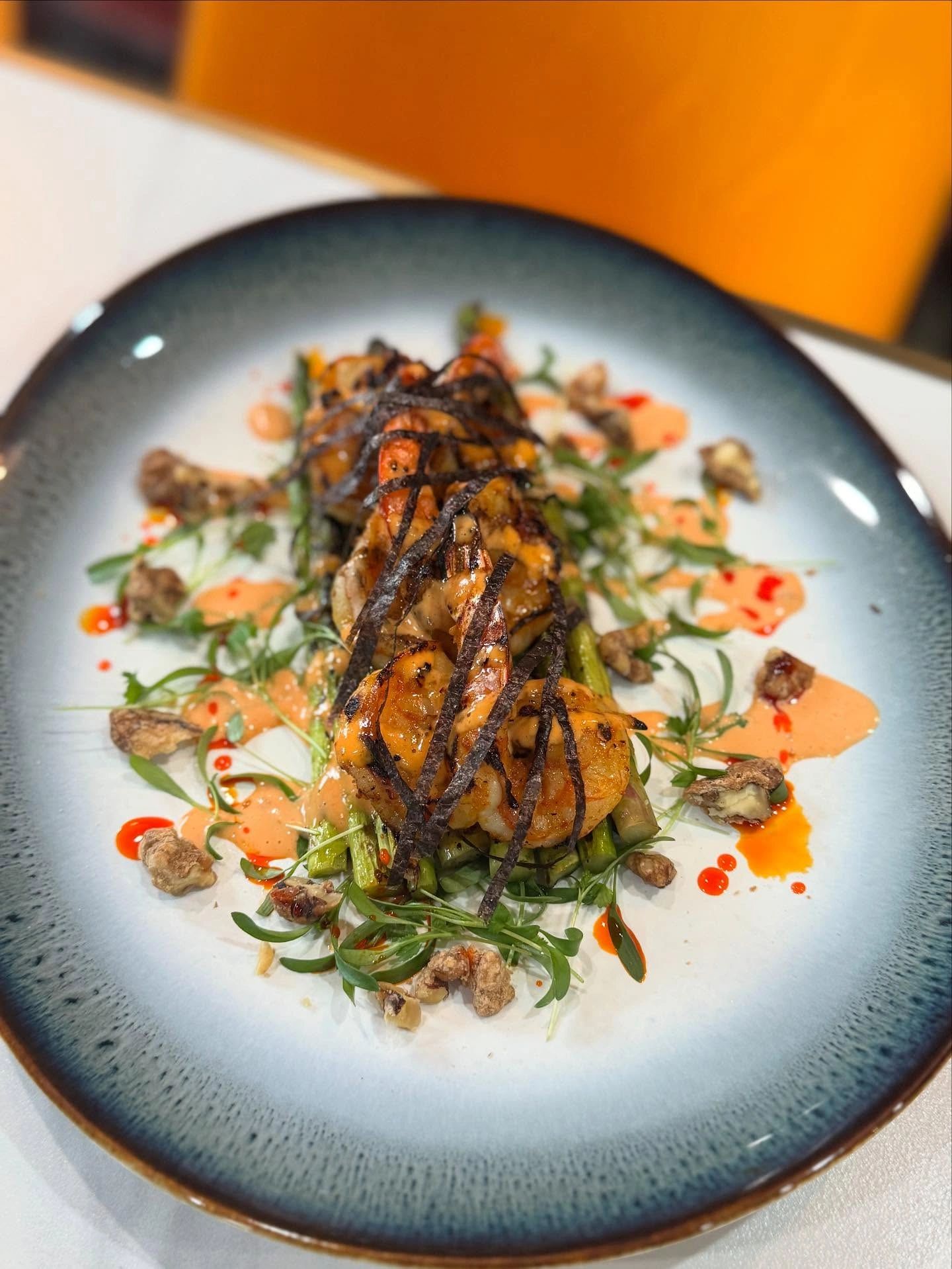 Grilled shrimp with asparagus, sauce, nuts, and herbs on a blue-edged plate.