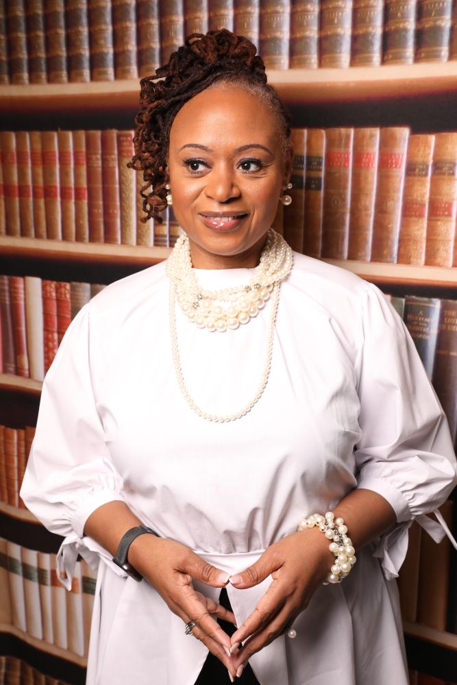 Woman in white blouse with pearls and dreadlocks posing against a bookcase backdrop.