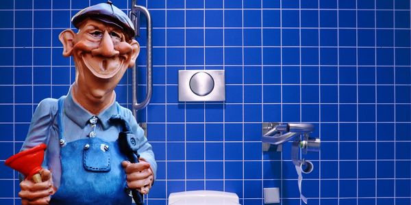 Pompel plumber figure holding tools and standing in front of commode in a blue tile bathroom