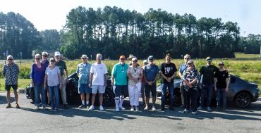 A group of people standing outdoors in front of cars on a sunny day.
