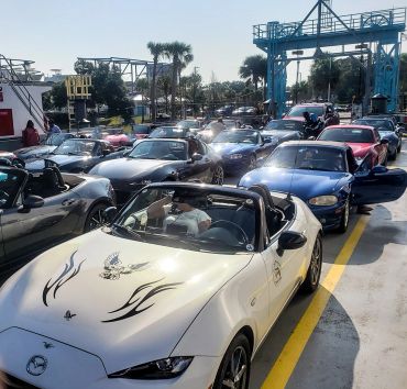 A line of Mazda Miata convertibles waiting on a ferry under clear skies.