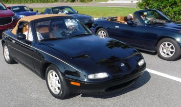 Two black Mazda Miata convertibles parked side by side with people inside.