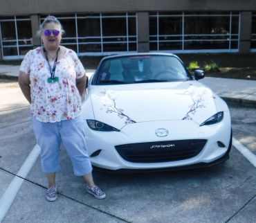 Person with sunglasses stands next to a white Mazda convertible with floral decals.