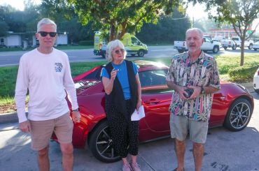 Three adults stand near a red sports car on a sunny day outdoors.