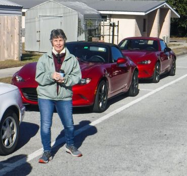 A smiling woman holding a coffee cup stands in front of two red sports cars on a sunny day.
