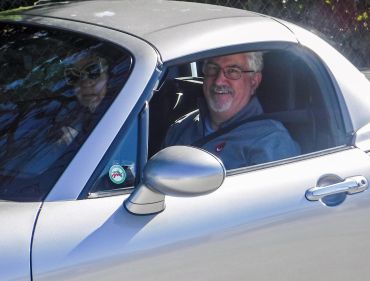 Two people inside a silver car, one smiling at the camera.