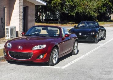 Two Mazda MX-5 Miata convertibles, one red and one black, parked in an outdoor lot.
