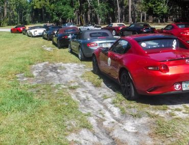 A line of Mazda MX-5 Miata cars parked on grass near a wooded area.