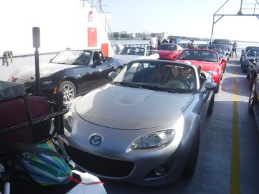 A line of Mazda convertible cars on a ferry with people standing in the background.