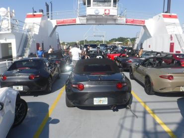 A group of Mazda MX-5 Miatas on a ferry with people standing around.